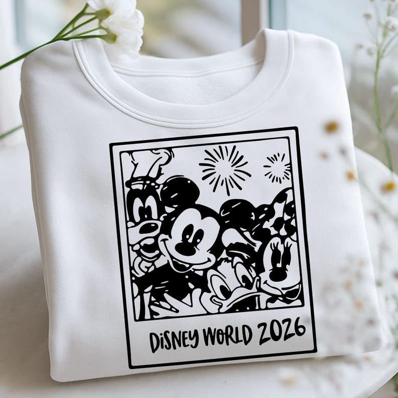 Op de afbeelding: Witte sweatshirt met een zwart-witte afbeelding van Disney-personages, waaronder Mickey Mouse, Goofy en Donald Duck. De afbeelding is omlijst als een foto met de tekst "DISNEY WORLD 2026". De sweatshirt heeft een ronde hals.