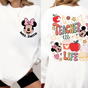 May include: White sweatshirt with a Minnie Mouse design, pink bow, and the name "Mrs. Smith" on the front. The back has the words "Teacher Life" with colourful graphics of apples, pencils, and Mickey Mouse.