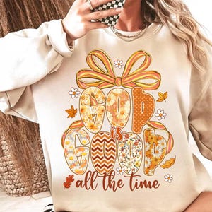 Puede incluir: Sudadera beige con las palabras "GOD IS GOOD" en letras grandes y coloridas. Las letras están decoradas con diseños florales y estampados, y la frase "all the time" está escrita debajo. Un gran lazo y hojas de otoño completan el diseño.