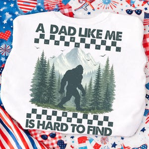 May include: White sweatshirt with the text "A DAD LIKE ME IS HARD TO FIND". The design features a silhouette of Bigfoot walking in front of a forest and mountains.