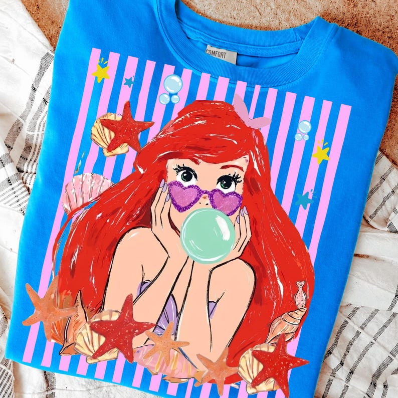May include: A blue sweatshirt with a cartoon mermaid graphic. The mermaid has red hair, heart-shaped sunglasses, and a bubble. The design includes seashells, starfish, and a pink and white striped background.
