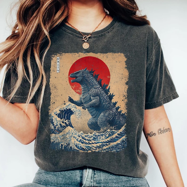 May include: Dark grey t-shirt with a Godzilla design in a Japanese art style. The graphic features a red sun, ocean waves, and the word "KAIJU". The design is printed on a vintage-style, washed fabric.