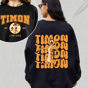 May include: Black sweatshirt with a graphic of Timon from The Lion King. The front of the sweatshirt has the text "Timon" with "Est. 1994" and "Lion King" underneath. The back of the sweatshirt has a repeated graphic of Timon with the text "Timon" in a retro font.