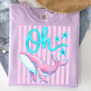 May include: A lilac-colored t-shirt featuring a pink and blue whale illustration with the word "Oh" in turquoise script. The design includes pink vertical stripes and turquoise stars.
