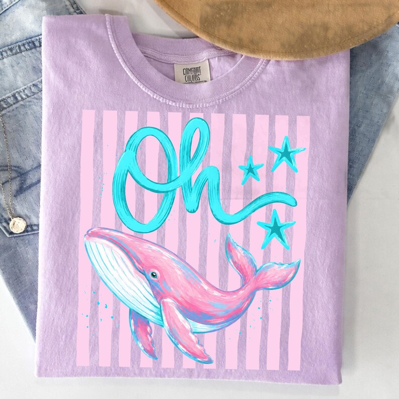 May include: A lilac-colored t-shirt featuring a pink and blue whale illustration with the word "Oh" in turquoise script. The design includes pink vertical stripes and turquoise stars.