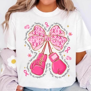 May include: White t-shirt featuring a pink bow with pink ribbon patterns and boxing gloves. The words "Together We Fight, Together We Win" are printed around the bow and gloves. The design includes pink ribbons, butterflies, and flowers.