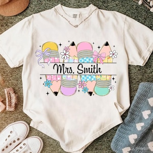 May include: Cream-colored t-shirt featuring a colorful pencil design with bows and flowers. The text "Mrs. Smith" is printed in black. The pencils are in shades of yellow, pink, and blue. The shirt has a round neckline.