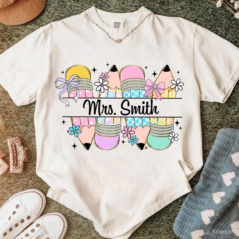 May include: Cream-colored t-shirt featuring a colorful pencil design with bows and flowers. The text "Mrs. Smith" is printed in black. The pencils are in shades of yellow, pink, and blue. The shirt has a round neckline.