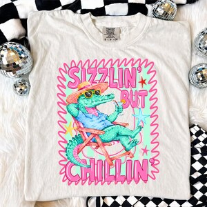 May include: Cream-colored t-shirt featuring a cartoon alligator in sunglasses, a hat, and a blue shirt, relaxing in a beach chair with a cocktail. The shirt has the text "Sizzlin' But Chillin'" in pink and green.