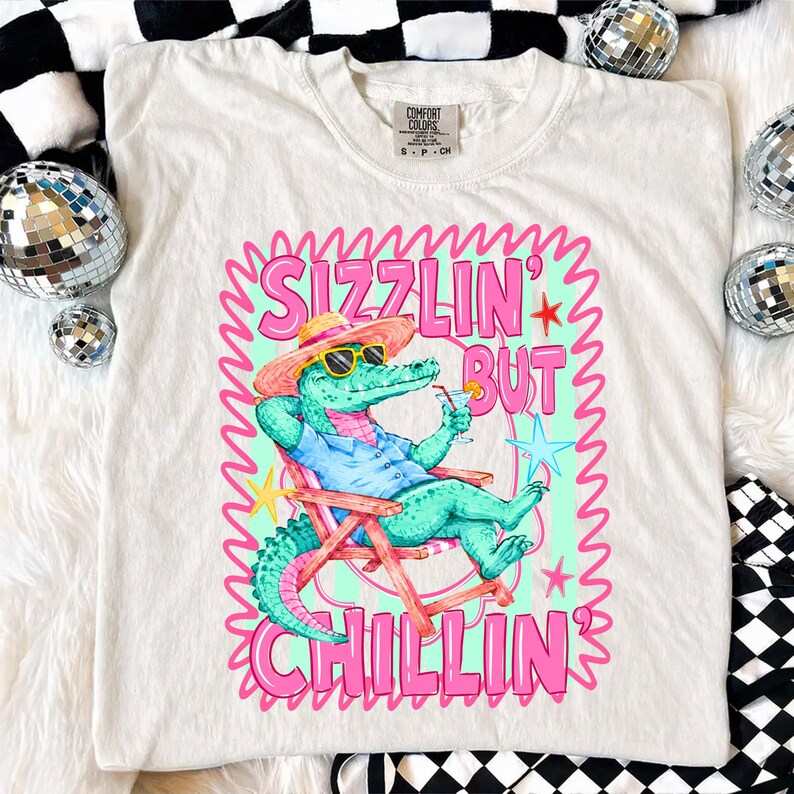 May include: Cream-colored t-shirt featuring a cartoon alligator in sunglasses, a hat, and a blue shirt, relaxing in a beach chair with a cocktail. The shirt has the text "Sizzlin' But Chillin'" in pink and green.