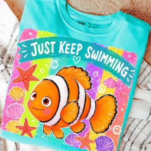 K&ouml;nnte beinhalten: Ein t&uuml;rkisfarbenes Sweatshirt mit Rundhalsausschnitt und einer farbenfrohen Grafik mit einem orange-wei&szlig; gestreiften Clownfisch, Seesternen und Muscheln. Der Text "JUST KEEP SWIMMING" ist &uuml;ber dem Fisch zu sehen. Das Design hat einen Regenbogenhintergrund.