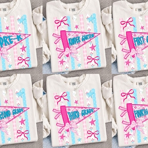 May include: Cream-colored sweatshirt with a school pennant design. The pennant is pink and features the grade level, such as "Pre-K" or "First Grade." The design includes pink bows, stars, and blue and pink striped accents.