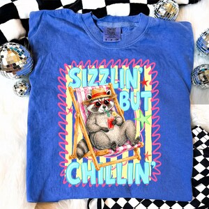 May include: Blue Comfort Colors t-shirt with a graphic of a raccoon in sunglasses relaxing in a beach chair, holding a drink. The text reads "Sizzlin' But Chillin'". The shirt has a crew neck and short sleeves.