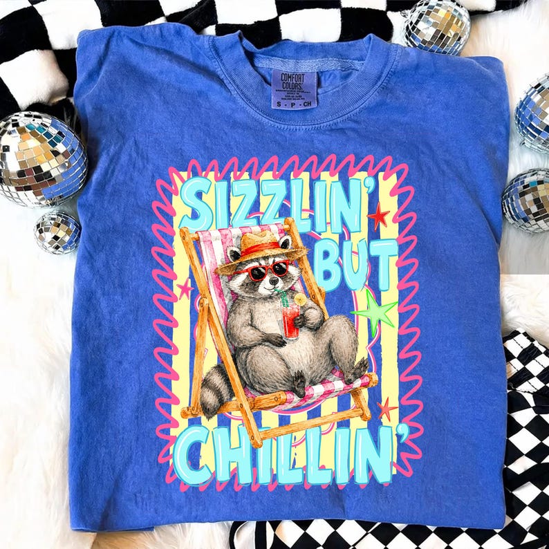 May include: Blue Comfort Colors t-shirt with a graphic of a raccoon in sunglasses relaxing in a beach chair, holding a drink. The text reads "Sizzlin' But Chillin'". The shirt has a crew neck and short sleeves.