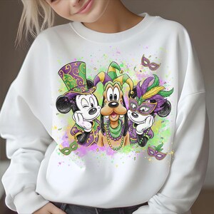 May include: White sweatshirt featuring Mickey Mouse, Goofy, and Minnie Mouse in Mardi Gras attire. Mickey Mouse wears a purple and green top hat, Goofy has a jester's hat, and Minnie Mouse wears a feathered mask. The design includes confetti and masks.