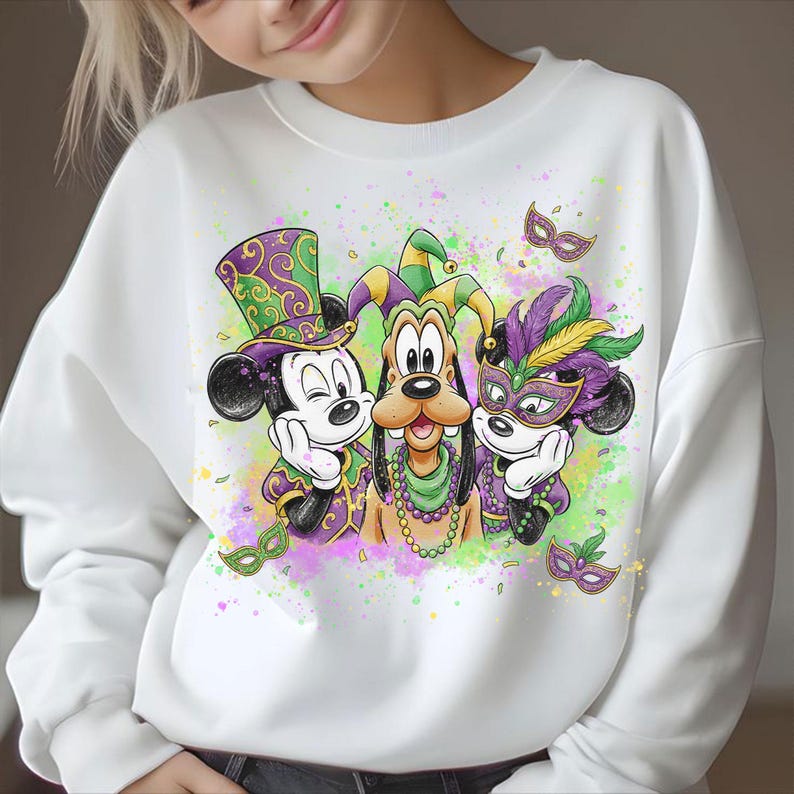 May include: White sweatshirt featuring Mickey Mouse, Goofy, and Minnie Mouse in Mardi Gras attire. Mickey Mouse wears a purple and green top hat, Goofy has a jester's hat, and Minnie Mouse wears a feathered mask. The design includes confetti and masks.