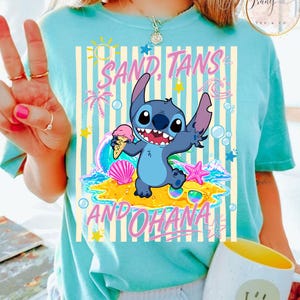 May include: Light blue t-shirt featuring a cartoon character holding ice cream on a beach with the words "SAND, TANS AND OHANA". The design includes a cartoon wave, starfish, and sunglasses.