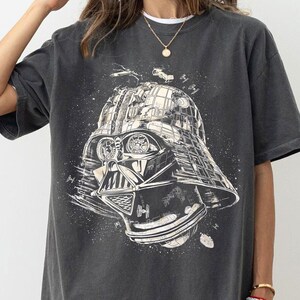 May include: Dark gray t-shirt featuring a detailed illustration of Darth Vader's helmet and face, with space battle elements. The design is in white and gray tones, creating a vintage, distressed look. The shirt is a crew neck.