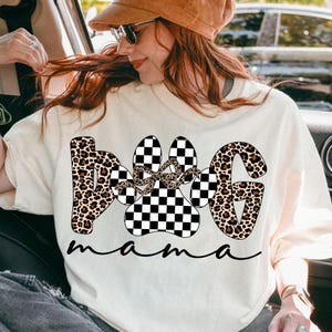 May include: Cream-colored t-shirt with the words "Dog Mama" in a mix of leopard print and checkered patterns. The paw print design features a bow. The shirt is a casual style.