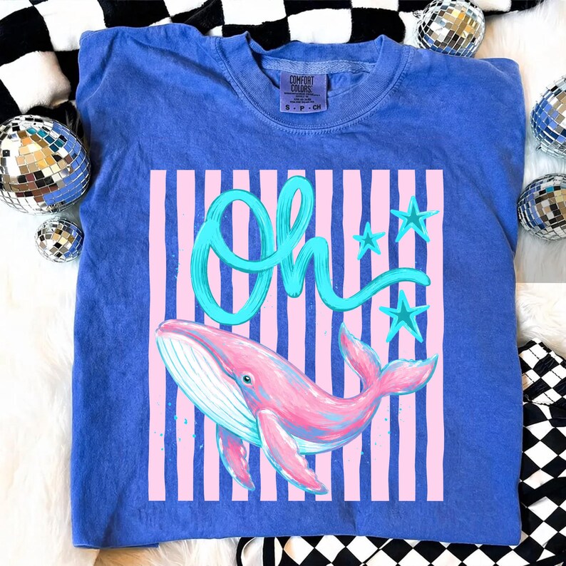 May include: A blue t-shirt with a pink and blue whale graphic. The shirt features the word "Oh" in teal script, along with teal stars and pink vertical stripes. The shirt is made of a soft material.
