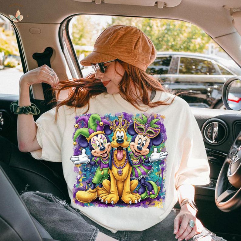May include: Cream-colored t-shirt featuring a colorful graphic of Mickey and Minnie Mouse, and Pluto, dressed in Mardi Gras attire. The design includes purple, green, and gold accents, with a vibrant, abstract background. The characters are wearing festive masks and crowns.