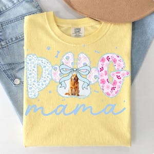 May include: Yellow t-shirt with the words "Dog Mama" in light blue script. The word "Dog" is designed with a floral "D", a paw print with a bow, and a floral "G". A golden retriever is in the paw print.