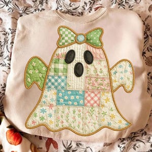 May include: A light pink sweatshirt featuring a patchwork ghost design. The ghost has a green and white checkered bow, black oval eyes, and a patchwork body with floral and checkered patterns. The design is outlined with a gold-colored trim.