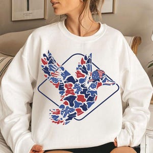 May include: White sweatshirt featuring a patriotic duck design. The duck is rendered in red, white, and blue camouflage patterns, wearing a baseball cap with an American flag design, and enclosed within a blue diamond frame.