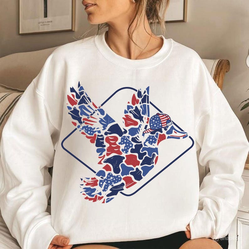 May include: White sweatshirt featuring a patriotic duck design. The duck is rendered in red, white, and blue camouflage patterns, wearing a baseball cap with an American flag design, and enclosed within a blue diamond frame.