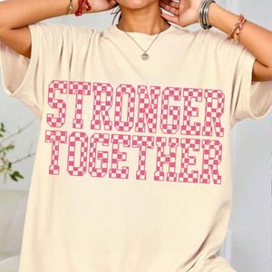 May include: Cream-colored t-shirt with the words "STRONGER TOGETHER" in a pink checkered pattern. The shirt has short sleeves and a relaxed fit. The text is large and centered on the front of the shirt.
