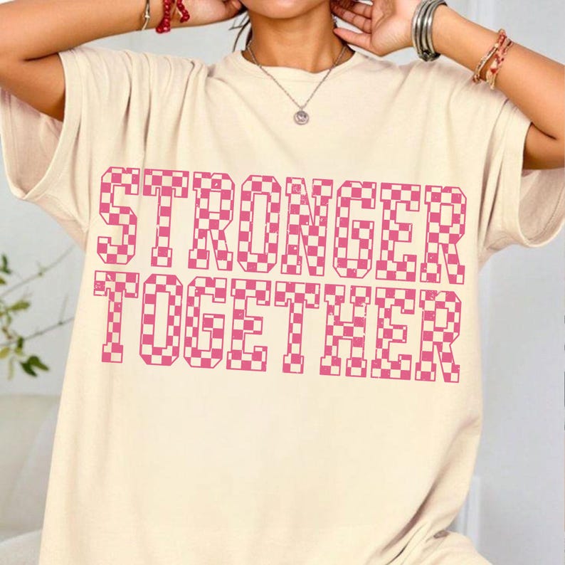 May include: Cream-colored t-shirt with the words "STRONGER TOGETHER" in a pink checkered pattern. The shirt has short sleeves and a relaxed fit. The text is large and centered on the front of the shirt.