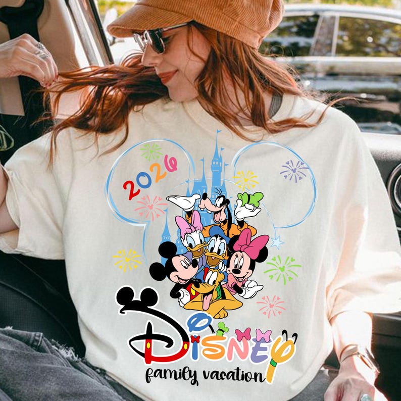 Może przedstawiać: Kremowa bluza z kolorowym wzorem wakacji rodzinnych Disneya. Grafika zawiera Myszkę Miki i Minnie, Kaczora Donalda i Daisy, Goofy'ego i Pluto. Rok "2026" jest wyświetlany nad postaciami, ze słowami "Disney" i "wakacje rodzinne" poniżej.
