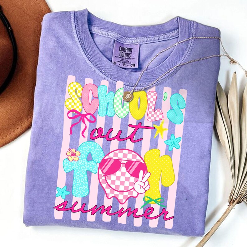 May include: A lilac Comfort Colors t-shirt with the words "School's Out For Summer" in colorful, playful lettering. The design includes a checkered ghost wearing sunglasses, stars, and a peace sign. A gold necklace is draped across the shirt.