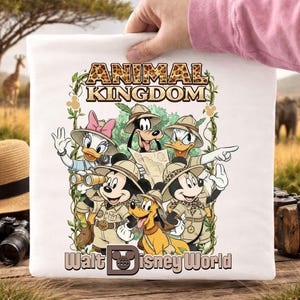 May include: White t-shirt featuring a colorful graphic of Disney characters in safari attire, including Mickey and Minnie Mouse, Donald and Daisy Duck, Goofy, and Pluto. The text "ANIMAL KINGDOM" and "Walt Disney World" are also displayed.