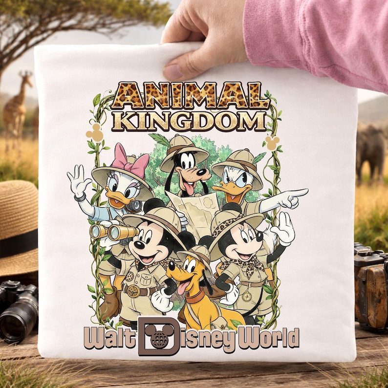 May include: White t-shirt featuring a colorful graphic of Disney characters in safari attire, including Mickey and Minnie Mouse, Donald and Daisy Duck, Goofy, and Pluto. The text "ANIMAL KINGDOM" and "Walt Disney World" are also displayed.