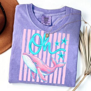 May include: A lilac Comfort Colors t-shirt featuring a pink whale illustration with blue accents and the word "Oh" in turquoise script. Pink and white vertical stripes are behind the whale. A gold necklace is draped across the shirt.