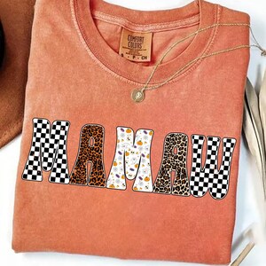 May include: An orange Comfort Colors t-shirt with the word "MAMA" in a retro font. The letters have different patterns: checkered, leopard print, and Halloween-themed. A gold necklace with a pendant is visible.