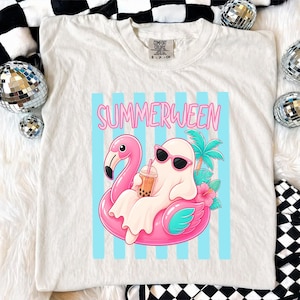 K&ouml;nnte beinhalten: Cremefarbenes T-Shirt mit einer Grafik eines Geistes mit Sonnenbrille, der auf einem rosa Flamingo-Schwimmring schwimmt. Der Geist h&auml;lt ein Getr&auml;nk. Das Wort "SUMMERWEEN" steht in rosa Buchstaben &uuml;ber der Grafik.