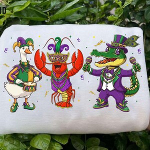 May include: White fabric featuring three cartoon characters in festive attire. A goose plays a drum, a lobster wears a mask, and an alligator holds maracas. All are dressed in purple, green, and gold, with confetti scattered around.