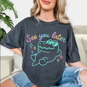 May include: Dark gray t-shirt with the phrase "See you later" in a colorful script. The shirt features a cartoon alligator wearing sunglasses and surrounded by stars. The shirt is a relaxed fit.