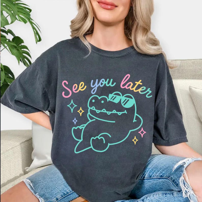 May include: Dark gray t-shirt with the phrase "See you later" in a colorful script. The shirt features a cartoon alligator wearing sunglasses and surrounded by stars. The shirt is a relaxed fit.