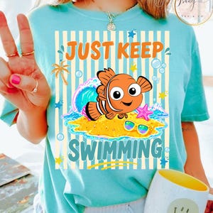 May include: Light blue t-shirt with a cartoon fish design. The design includes the text "JUST KEEP SWIMMING" with a cartoon fish, sunglasses, and a starfish on a sandy beach. The shirt has vertical stripes.