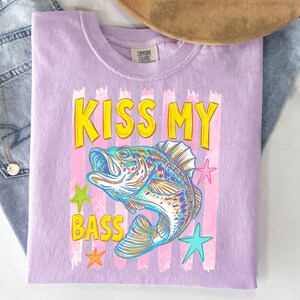May include: A light purple t-shirt with the text "KISS MY BASS" in yellow letters with a black outline. The shirt features a colorful illustration of a fish and star shapes.