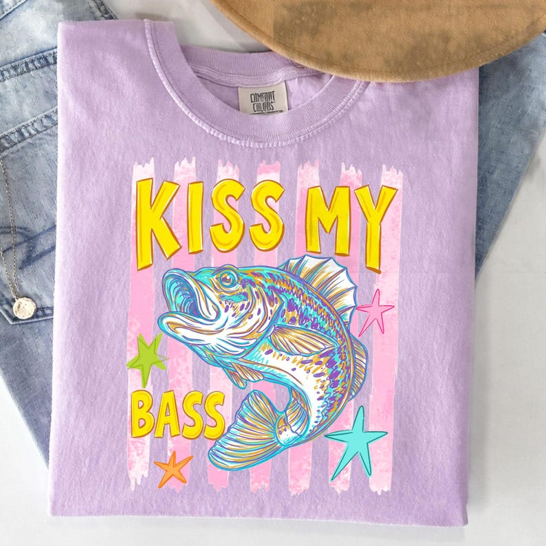 May include: A light purple t-shirt with the text "KISS MY BASS" in yellow letters with a black outline. The shirt features a colorful illustration of a fish and star shapes.