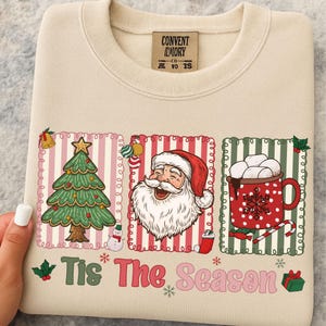 Puede incluir: Sudadera color crema con un dise&ntilde;o navide&ntilde;o festivo. El dise&ntilde;o incluye un &aacute;rbol de Navidad, Pap&aacute; Noel y una taza de chocolate caliente. El texto "Tis The Season" est&aacute; impreso en la parte inferior.