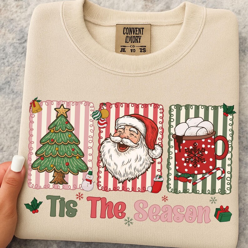Puede incluir: Sudadera color crema con un dise&ntilde;o navide&ntilde;o festivo. El dise&ntilde;o incluye un &aacute;rbol de Navidad, Pap&aacute; Noel y una taza de chocolate caliente. El texto "Tis The Season" est&aacute; impreso en la parte inferior.