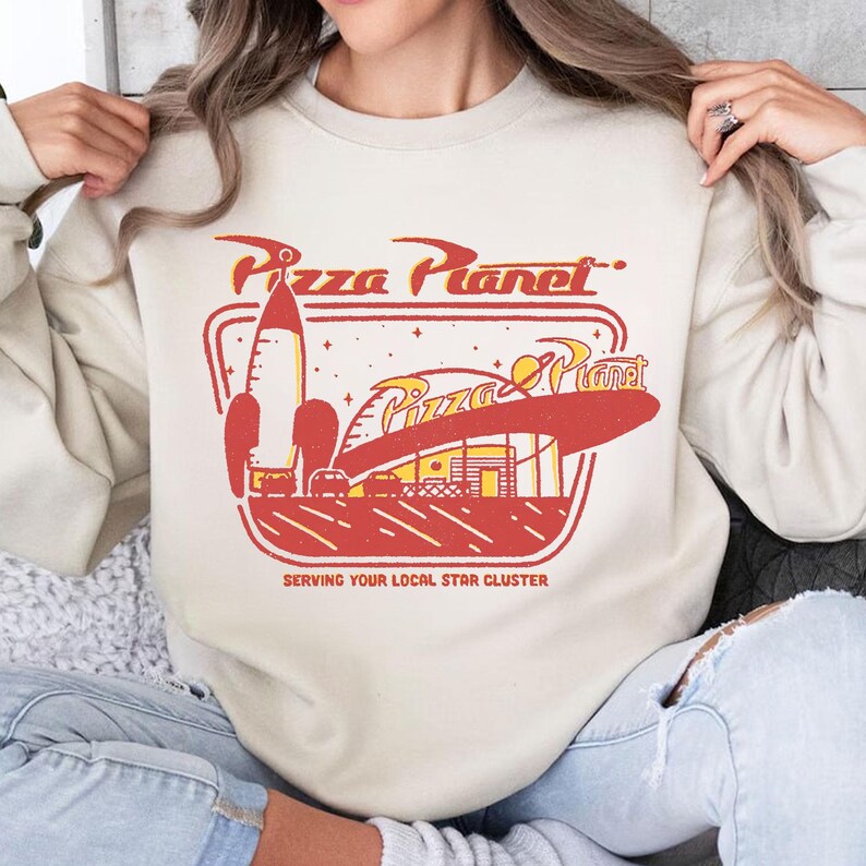 Puede incluir: Sudadera color crema con un gr&aacute;fico retro de Pizza Planet. El dise&ntilde;o incluye un cohete rojo, un logotipo estilizado de Pizza Planet y el texto "SERVING YOUR LOCAL STAR CLUSTER". Una prenda divertida de inspiraci&oacute;n vintage.