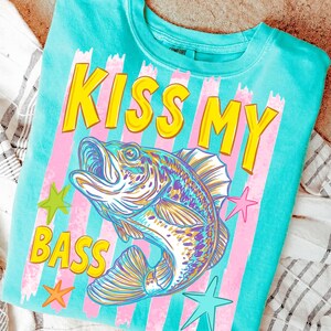 May include: A turquoise crew neck sweatshirt with the phrase "KISS MY BASS" in yellow block letters. A colorful illustration of a fish is centered below the text, with pink and white stripes and star accents.