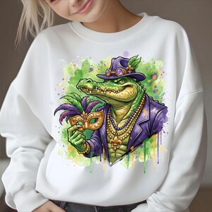 May include: White sweatshirt featuring a cartoon alligator in a purple suit, hat, and mask, with a beaded necklace. The alligator is green and yellow, set against a green and purple watercolor background. The design is a Mardi Gras theme.