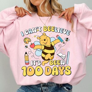 May include: Pink sweatshirt with a graphic featuring Winnie the Pooh as a bee, holding a pencil and apple. Text reads "I CAN'T BEELIEVE IT'S BEEN 100 DAYS" with school-themed illustrations. A fun, colorful design.